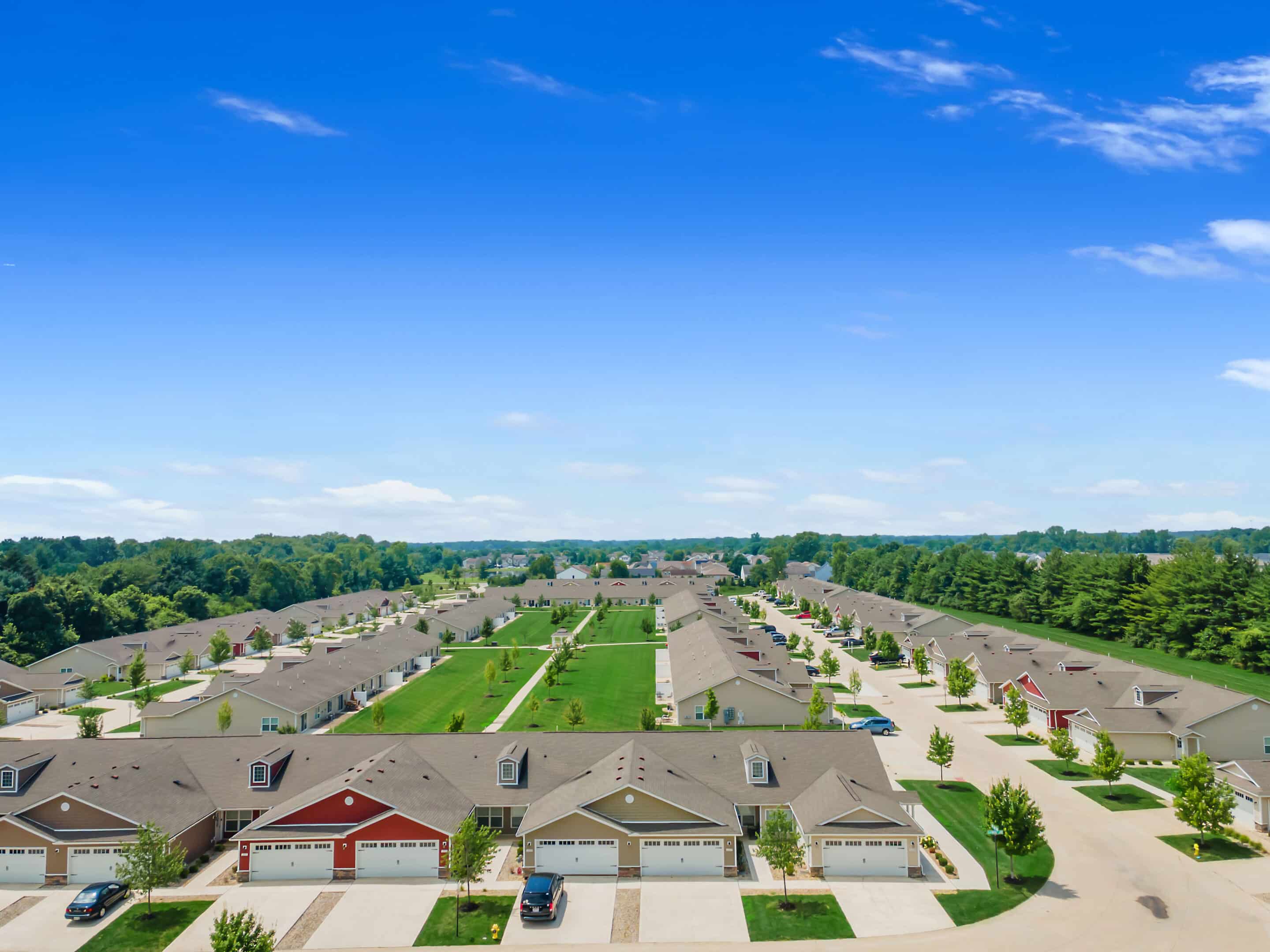 Aerial view of a suburban neighborhood with rows of single-story houses, green lawns, and a central grassy area under a blue sky.