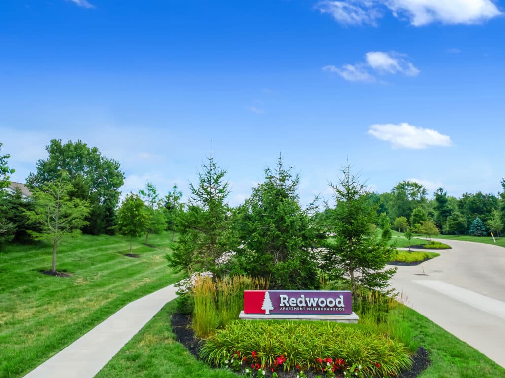 Redwood Apartments sign stands in landscaped greenery beside a wide driveway, with trees and neatly trimmed grass under a clear blue sky.