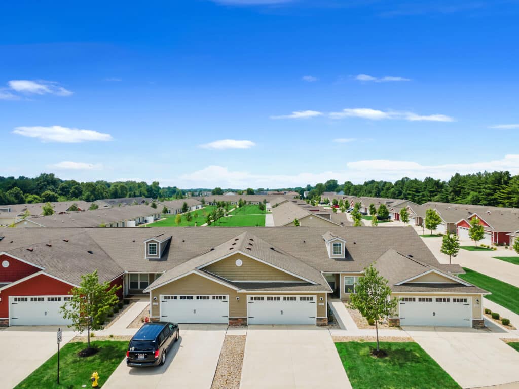 Aerial view of a suburban neighborhood with rows of identical single-family homes, driveways, lawns, and a parked black car in front of one house under a clear blue sky.