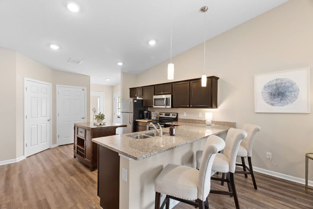 Modern kitchen with granite countertops, dark cabinets, stainless steel appliances, island with sink, and three upholstered barstools. Light beige walls and wood flooring throughout.