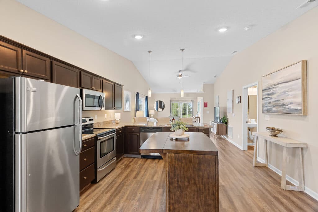 Modern kitchen with stainless steel appliances, dark wood cabinets, a central island, and adjacent open-plan living area with large windows and wood-style flooring.