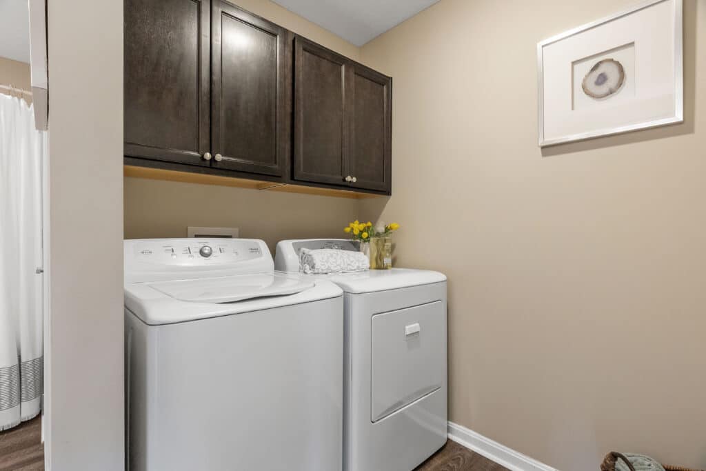 A laundry room with a white washing machine and dryer beneath dark wood cabinets, a decorative tray with towels, a vase with yellow flowers, and framed wall art.