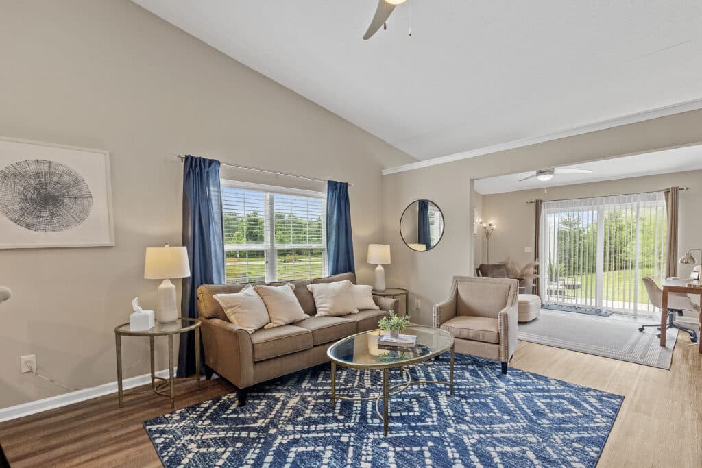 Bright living room with a beige sofa, armchair, round glass coffee table, blue patterned rug, large window with blue curtains, and sliding doors leading to a dining area.
