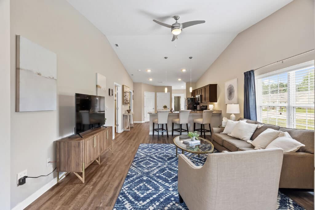 Open-concept living room and kitchen with neutral decor, a blue patterned rug, beige sofa, TV, dining area, and large windows with white blinds.