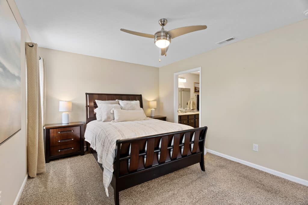 A bedroom with a dark wood bed, beige bedding, two matching nightstands with lamps, carpeted floor, and a ceiling fan; a bathroom is visible through an open doorway.