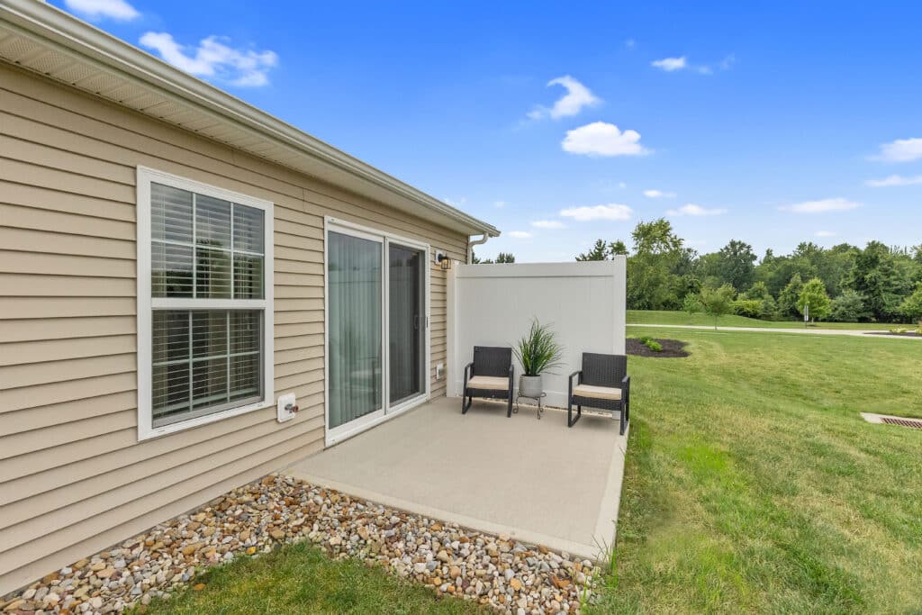 A small concrete patio with two black chairs, a potted plant, sliding glass door, and beige siding, overlooks a grassy yard with trees in the background.
