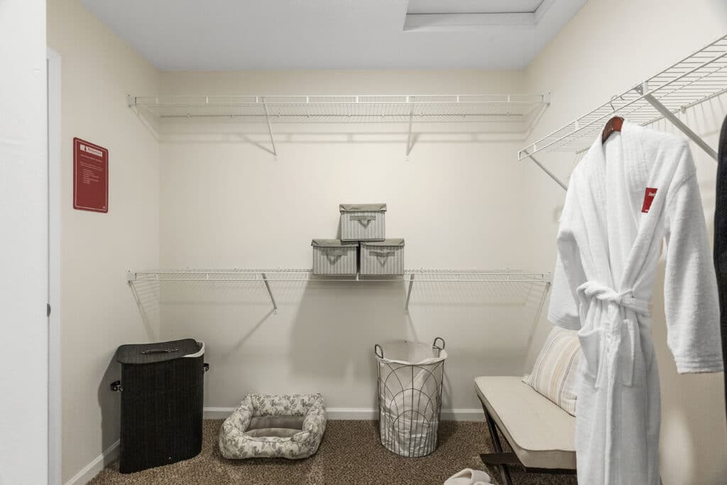 Spacious walk-in closet with wire shelving, storage baskets, a laundry hamper, a dog bed, a bench with pillows, and a white bathrobe hanging on the wall.