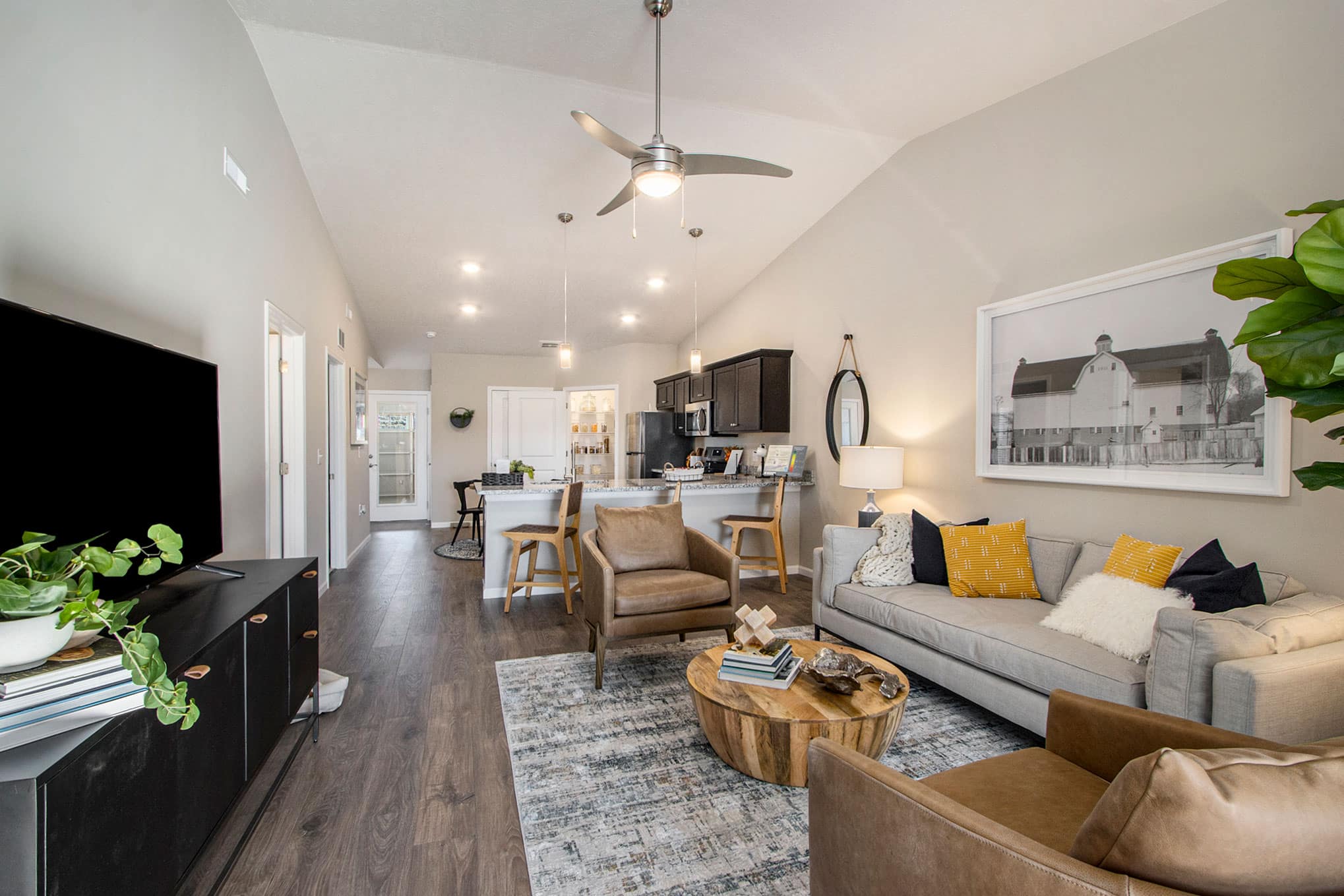 Modern open-concept living room and kitchen with neutral décor, wood flooring, a beige sofa, two brown chairs, and a large wall art above the couch.