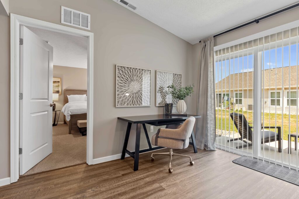A modern home office with a black desk, beige chair, decorative wall art, and sliding glass door leading to a patio with outdoor seating. A bedroom is visible through an open door.