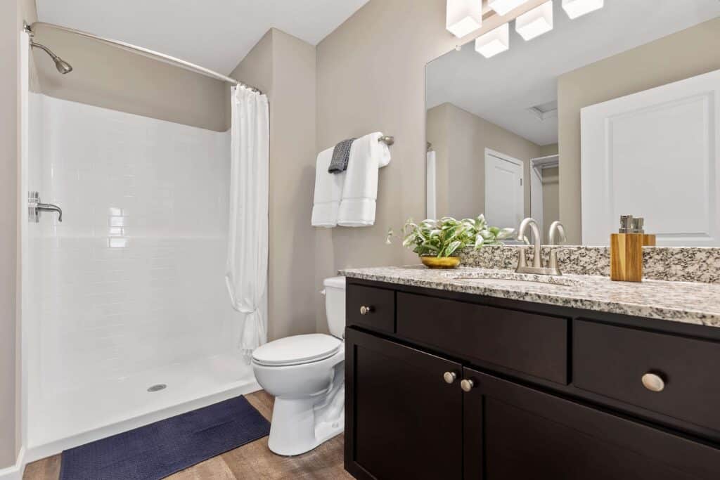 Modern bathroom with a glassless shower, white curtain, toilet, dark vanity with granite countertop, large mirror, and neatly folded towels on the wall.