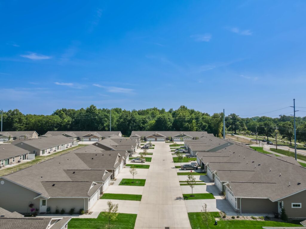 Aerial view of a suburban residential neighborhood with single-story houses, driveways, and a central concrete road, bordered by trees under a clear blue sky.