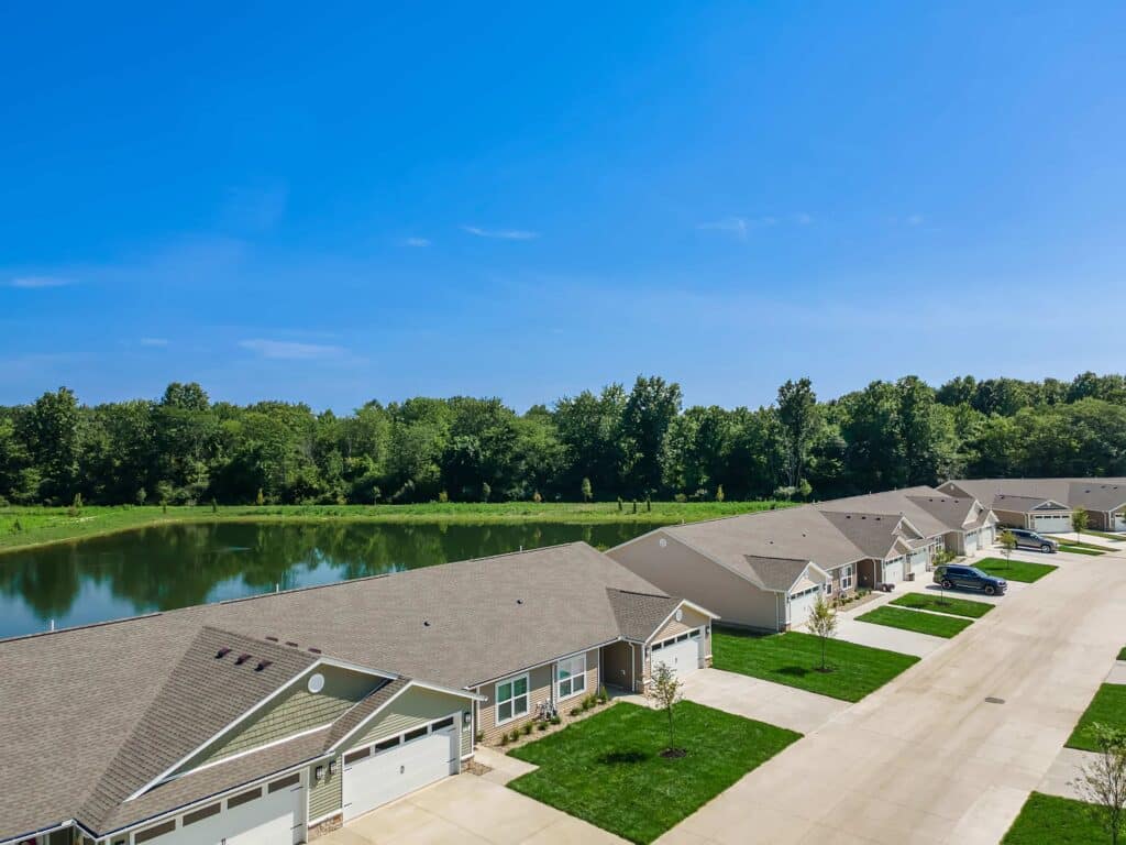 Row of single-story houses with attached garages facing a street, with a pond and trees in the background under a clear blue sky.