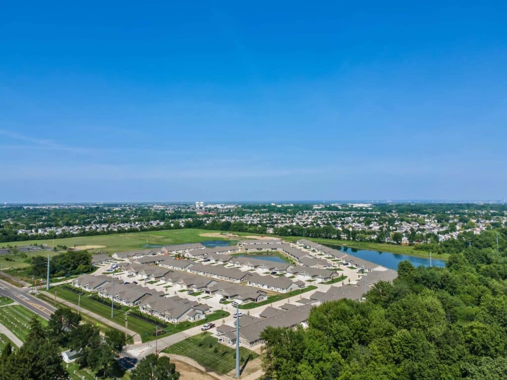 Aerial view of a suburban neighborhood with rows of houses, surrounding roads, green areas, and a small lake under a clear blue sky.