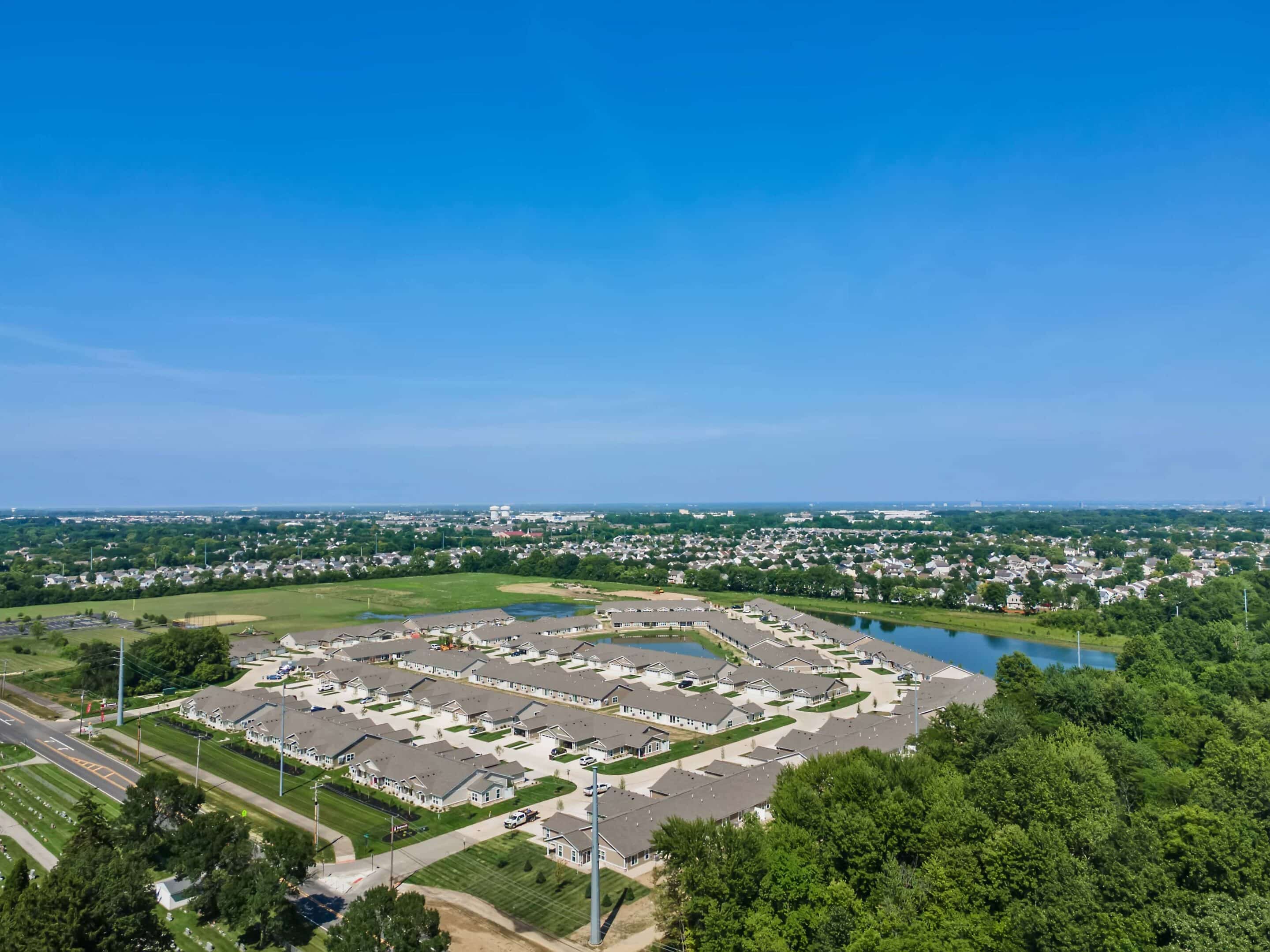 Aerial view of a suburban neighborhood with rows of houses, surrounding roads, green areas, and a small lake under a clear blue sky.