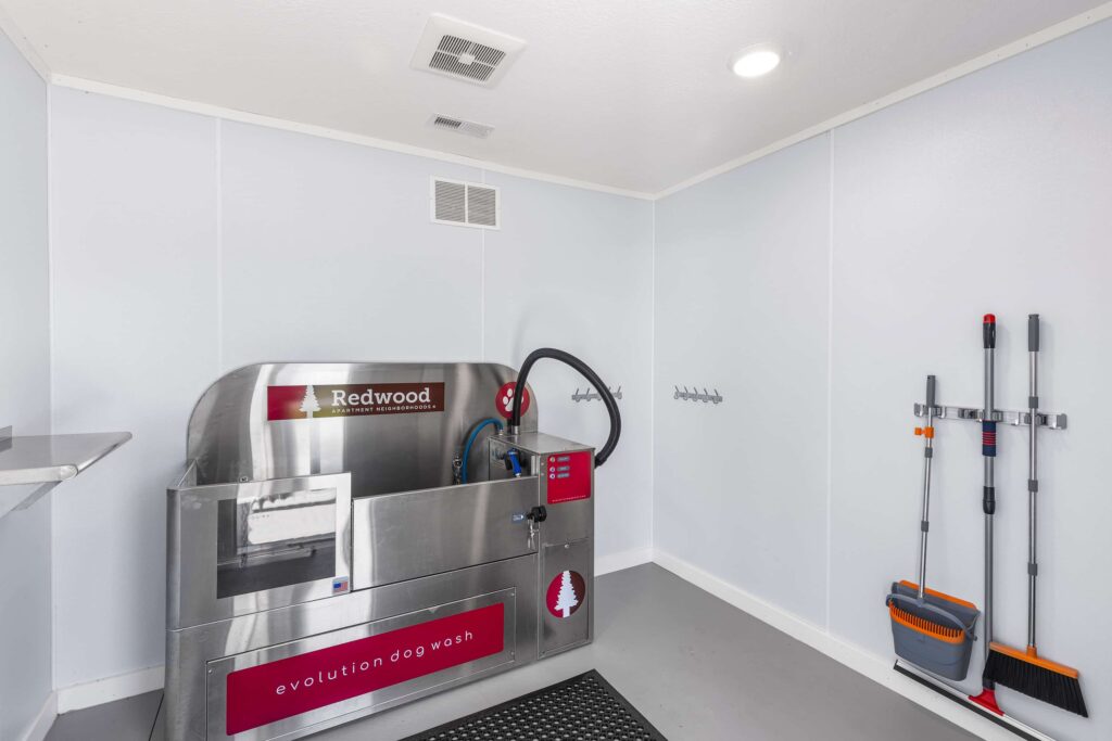 A stainless steel self-serve dog wash station in a clean, white room with hooks on the wall and cleaning supplies stored in the corner.