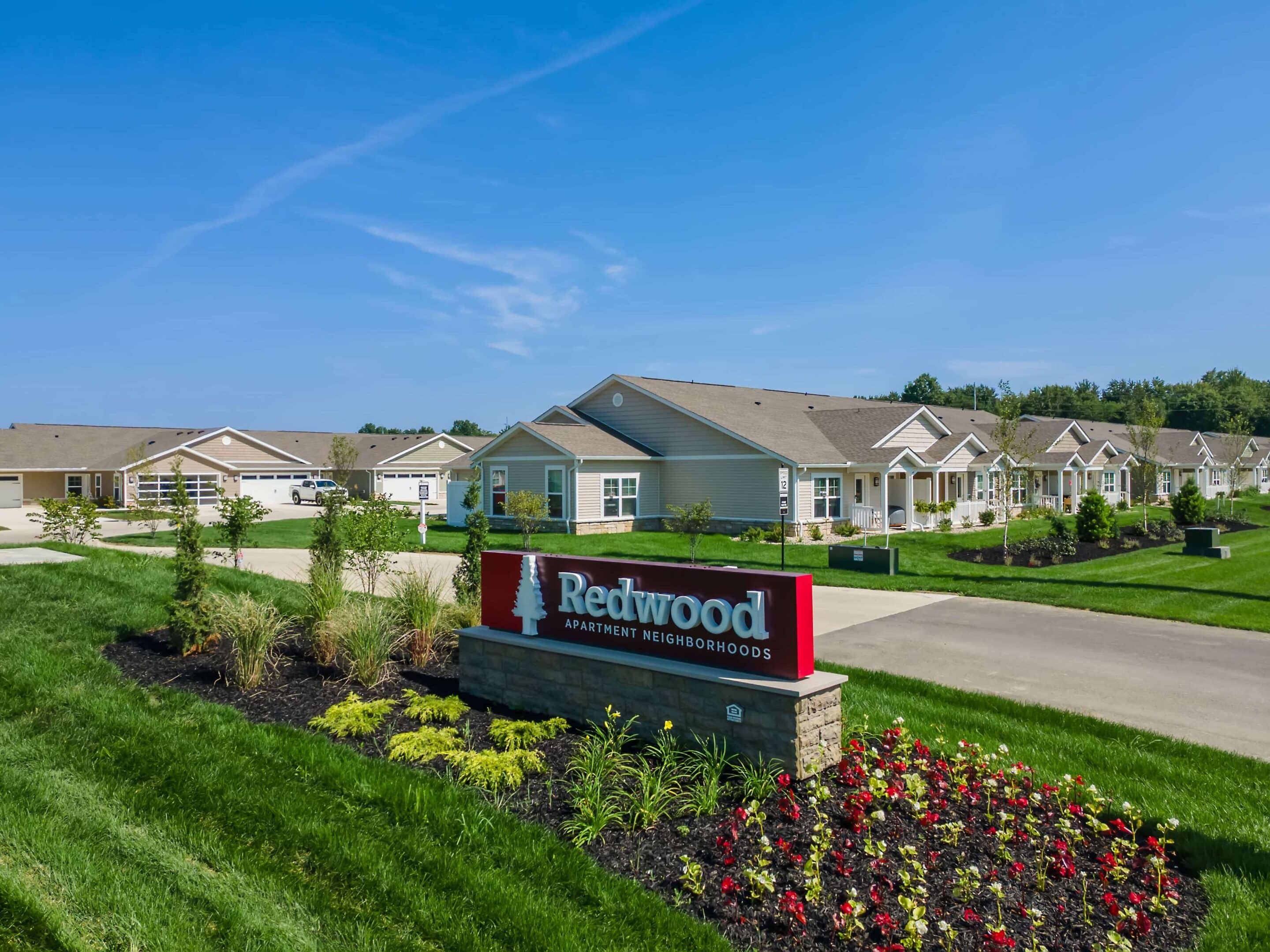Single-story apartment buildings with manicured lawns and landscaping, featuring a red sign that reads “Redwood Apartment Neighborhoods” in the foreground.