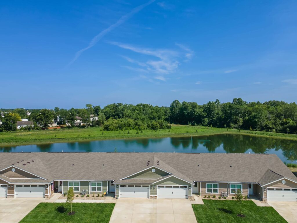 Row of single-story houses with attached garages facing a pond, with trees and blue sky in the background.