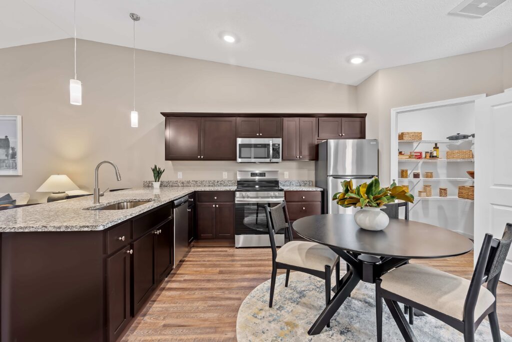 Modern kitchen with dark wood cabinets, stainless steel appliances, granite countertops, round dining table, and an open pantry with shelves in the background.