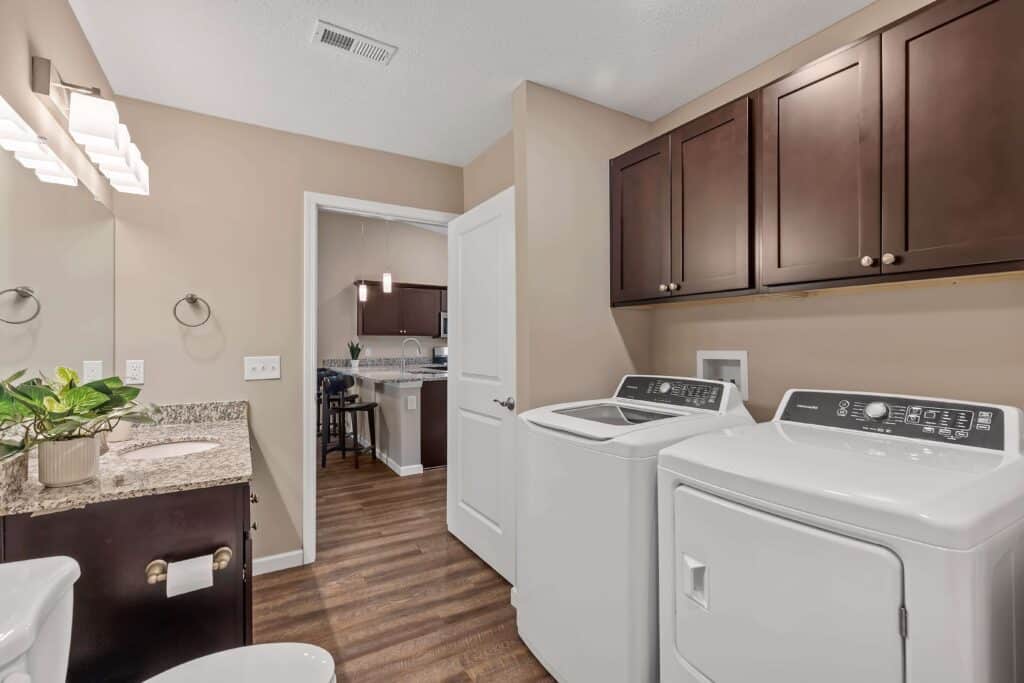 Laundry room with beige walls, dark wood cabinets, a washer and dryer, granite countertop with a sink, and a view into a kitchen area through an open door.