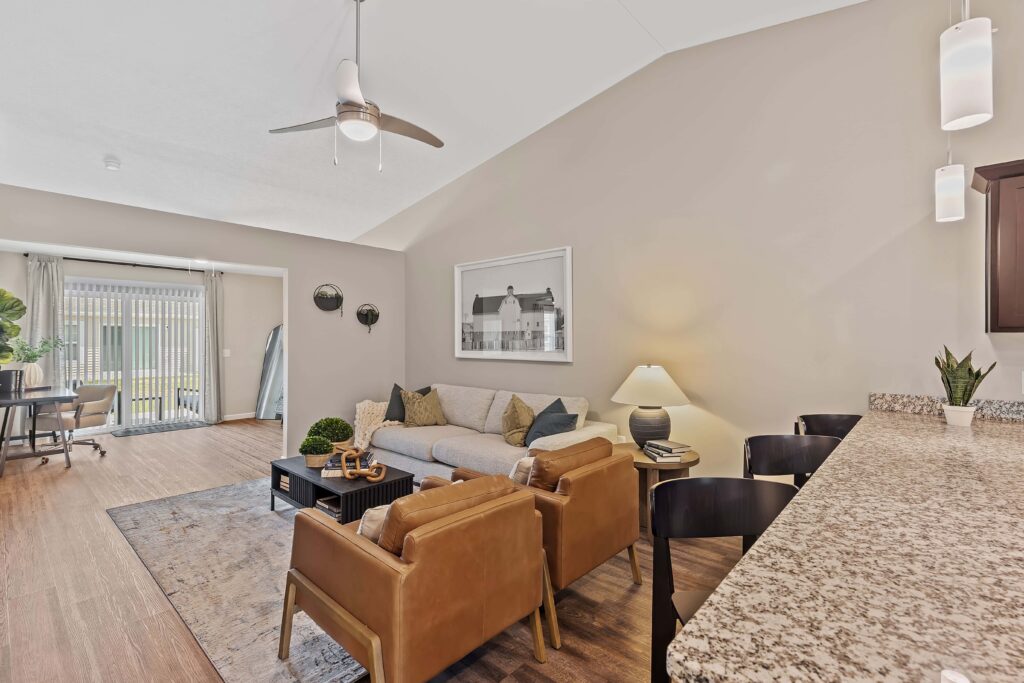 Modern living room with a gray sofa, two brown armchairs, a coffee table, and bar seating at a granite countertop. Sliding glass doors lead to a patio, and natural light fills the space.