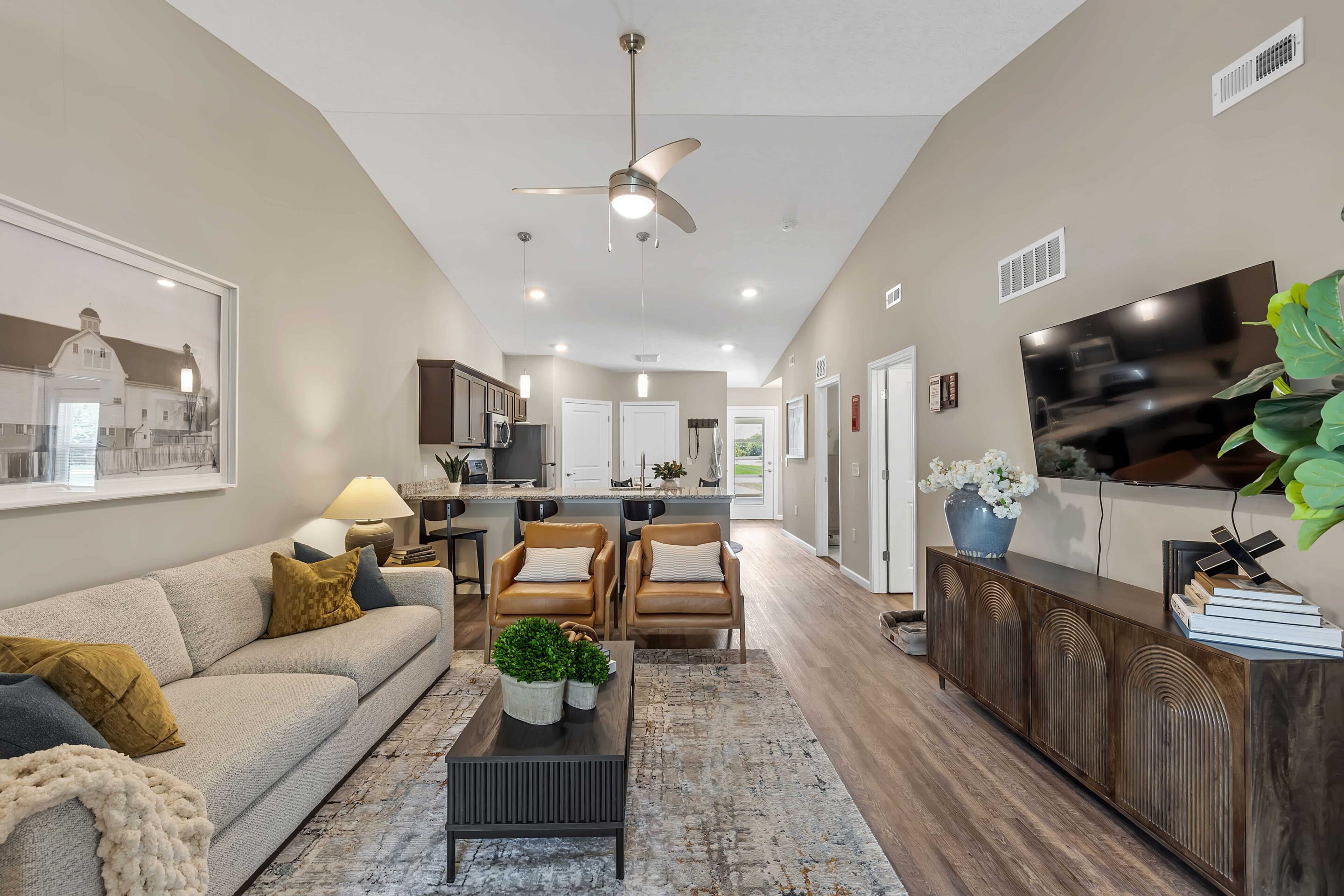 Modern living room with neutral colors, a sofa, two chairs, a rug, a wall-mounted TV, and an open view of the kitchen and hallway in the background.