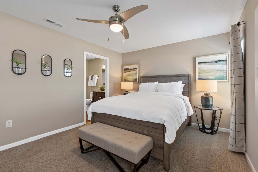 A bedroom with a wooden bed, white bedding, two side tables with lamps, a bench at the foot of the bed, beige walls, and a ceiling fan. An open door reveals a bathroom.