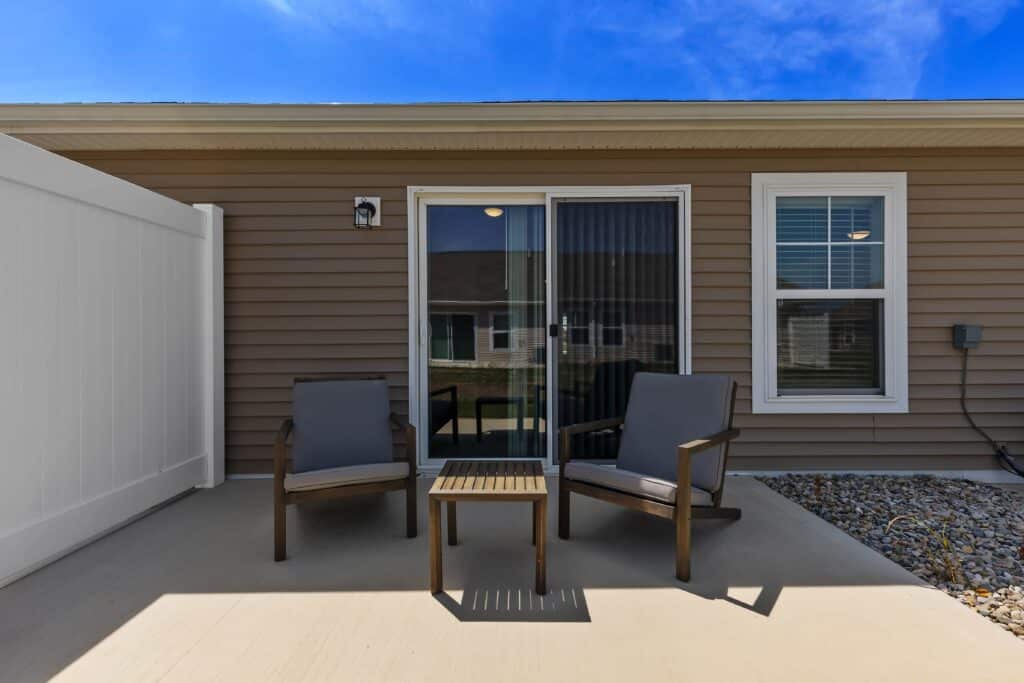 Two outdoor chairs and a small table on a concrete patio in front of a house with brown siding, a sliding glass door, and a window.