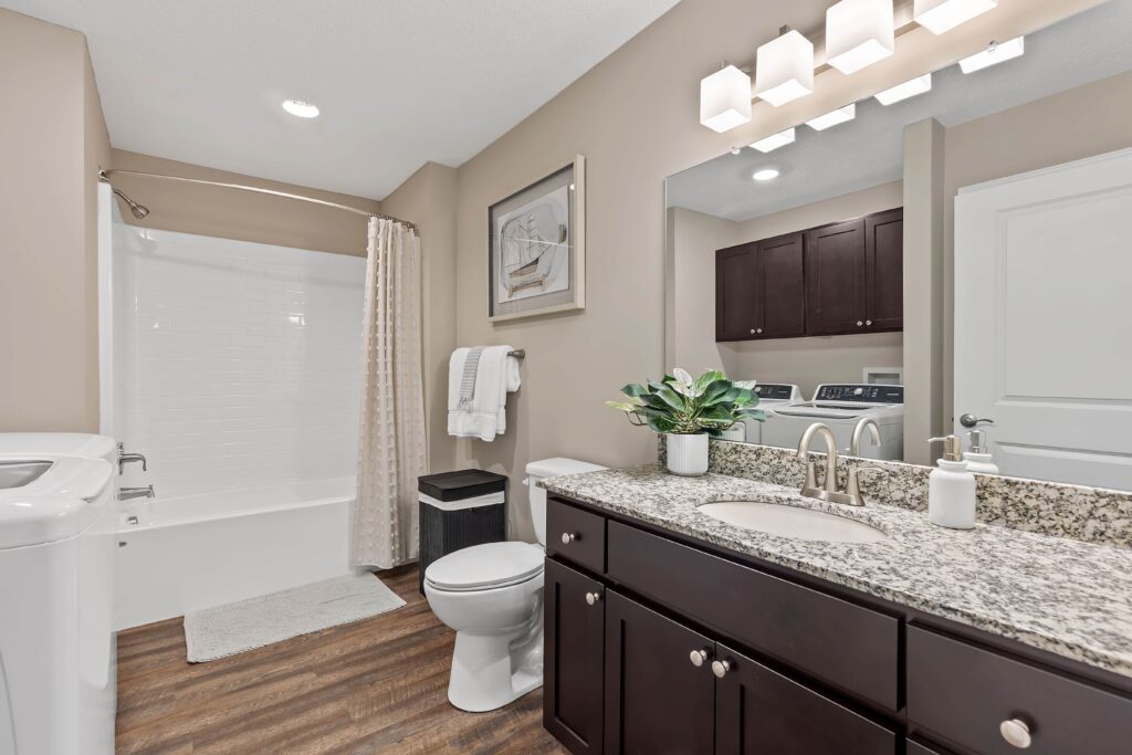 Modern bathroom with a granite countertop, dark wood cabinets, white bathtub with a curtain, washer and dryer, and a potted plant on the vanity.