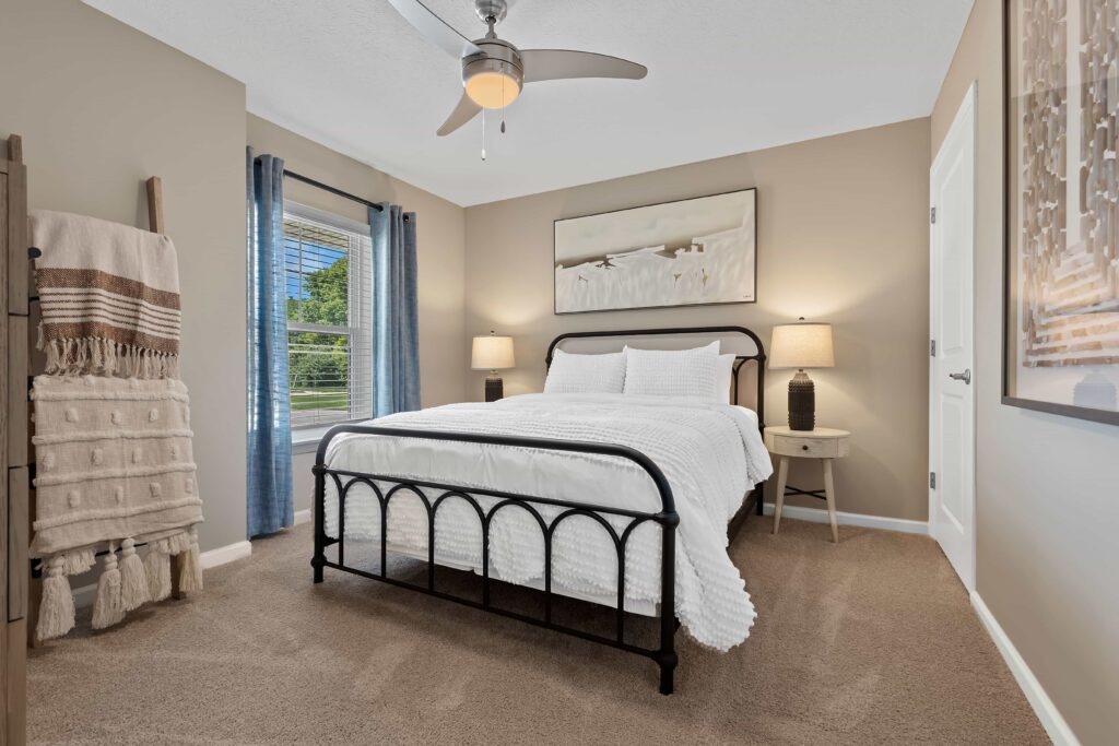 A neatly made bed with white bedding and a black metal frame is centered in a carpeted bedroom with beige walls, two nightstands, lamps, and artwork above the headboard.