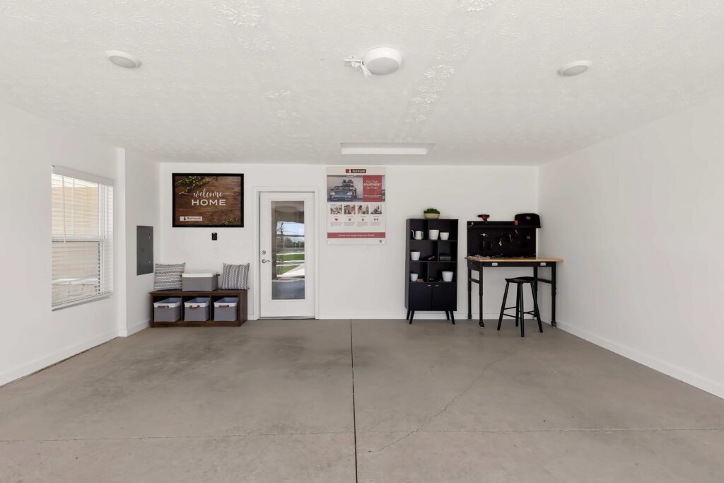 A clean, empty garage with white walls, minimal decor, a door with a window, storage bins, a black shelving unit, and a small bar table with two stools.