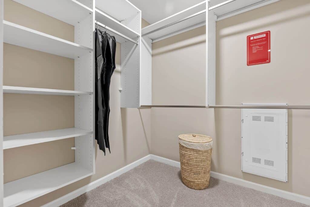 Empty walk-in closet with white shelving, a pair of black pants hanging, a wicker laundry basket on the carpeted floor, and a red sign on the wall.
