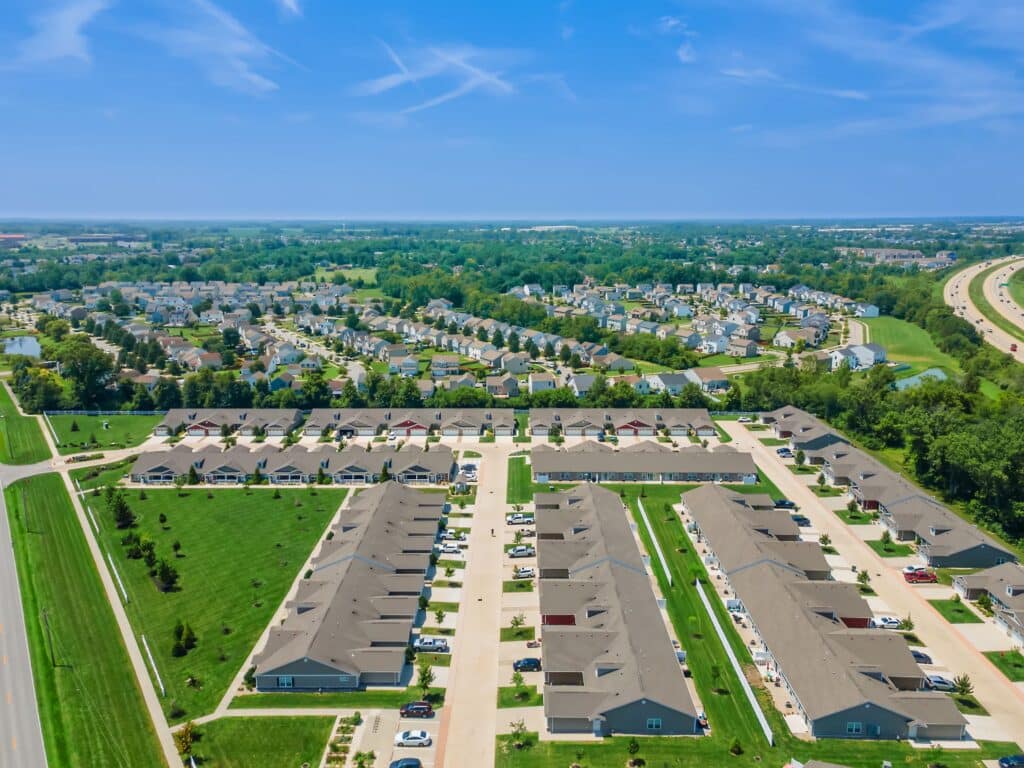 Aerial view of a suburban neighborhood with rows of houses, streets, green lawns, and trees, with a highway visible to the right.