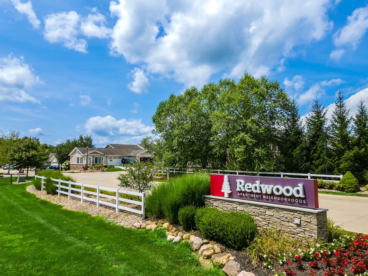 Entrance to Redwood Apartment Neighborhoods with a red sign, landscaped greenery, a white fence, and residential buildings in the background under a partly cloudy sky.