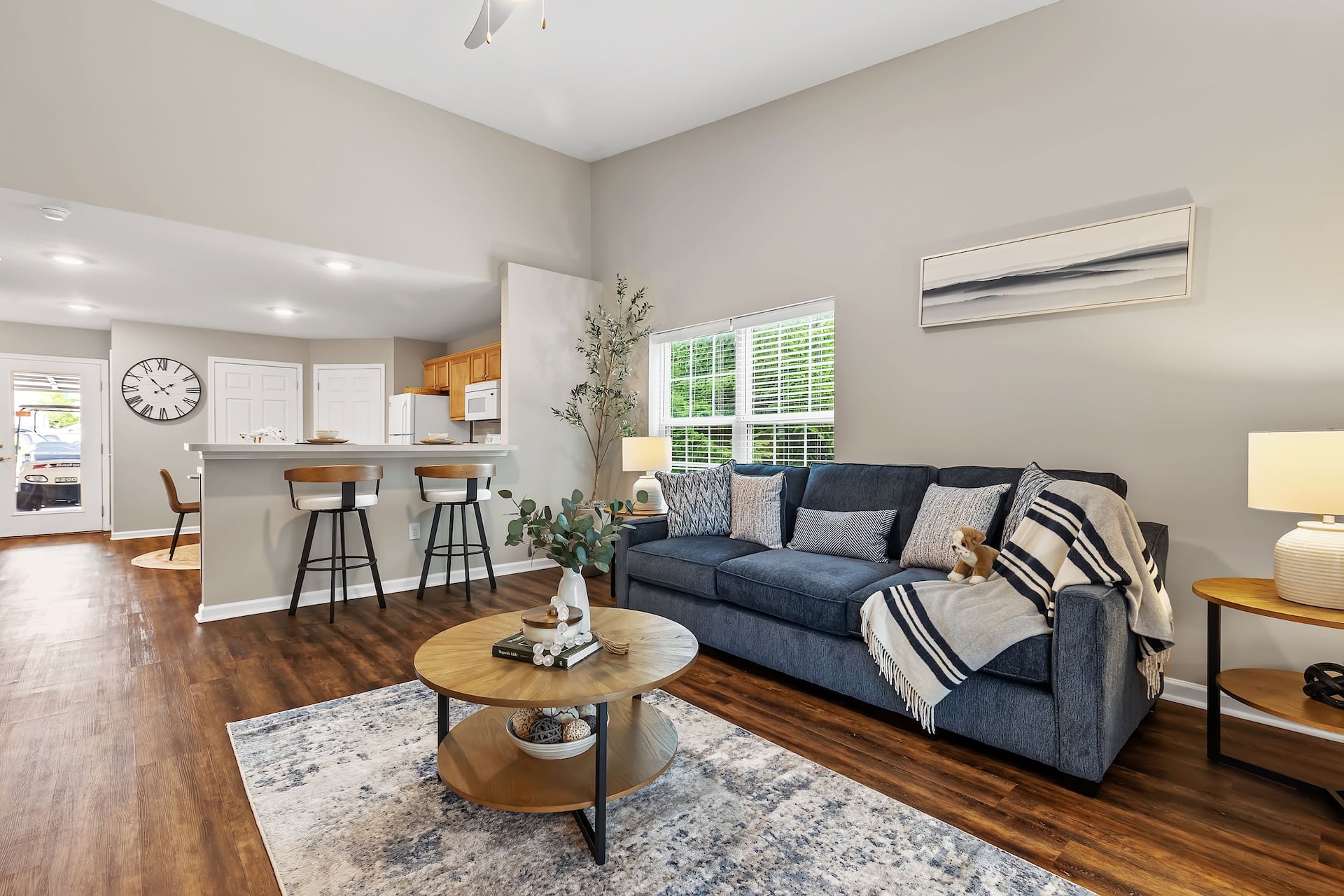 Modern living room with a blue sofa, round wooden coffee table, area rug, and open view to a kitchen with bar stools. The space features wood flooring and neutral decor.