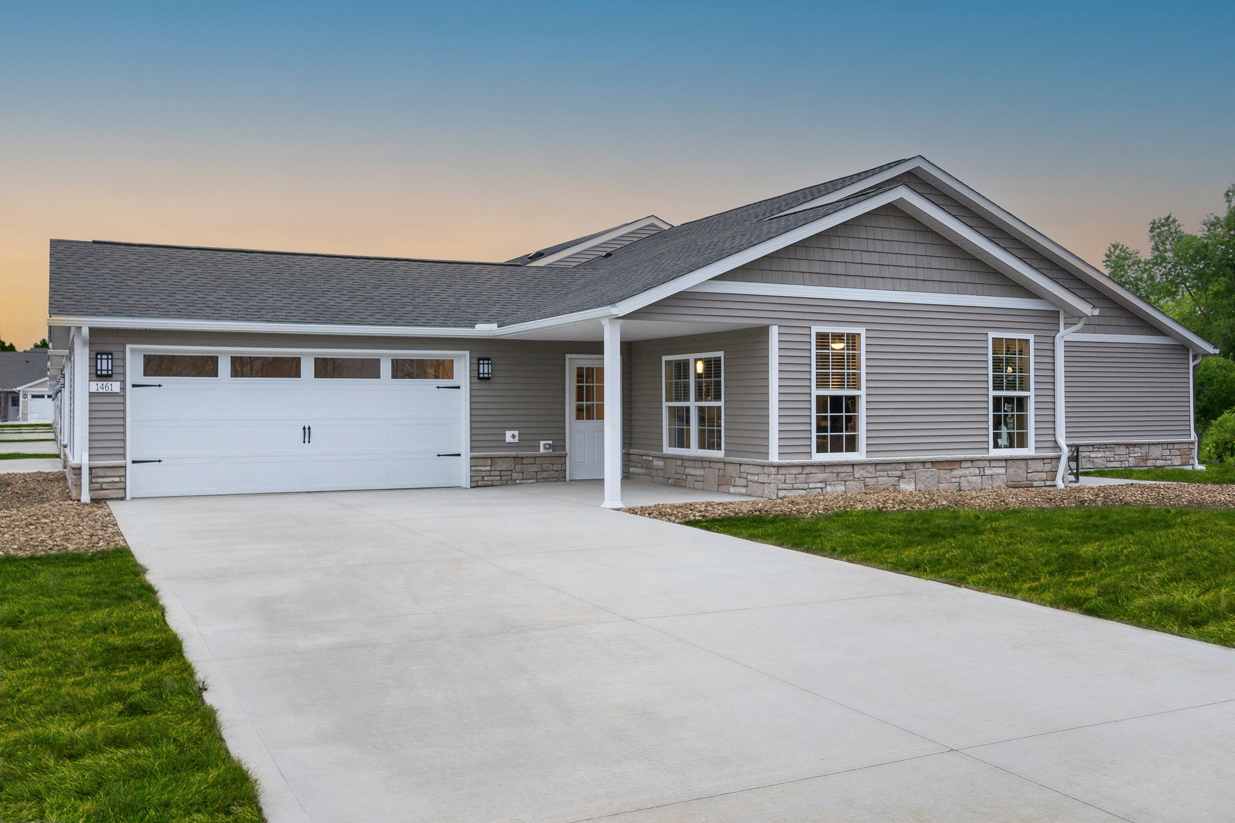 Single-story suburban house with gray siding, white trim, a two-car garage, front porch, and concrete driveway at sunset.