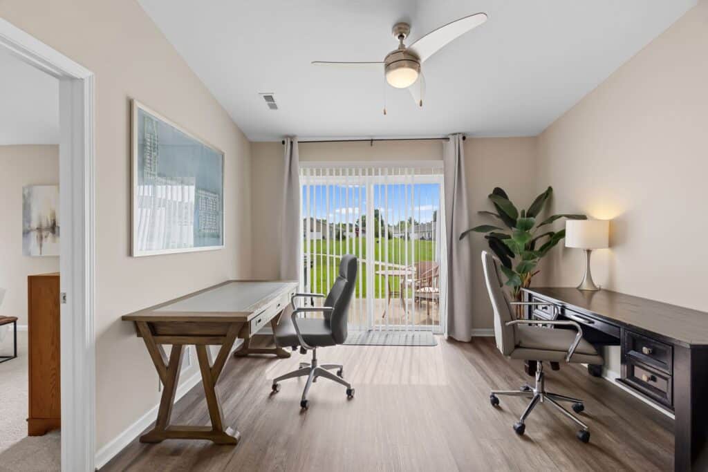 Modern home office with two desks, office chairs, a large plant, and a lamp, featuring sliding glass doors opening to a patio with a view of a green lawn.