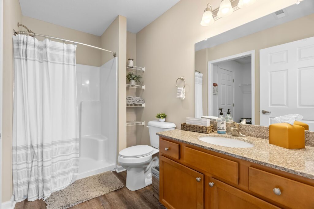 Bathroom with a shower-stall and curtain, toilet, shelves with towels, and a wooden vanity with a granite countertop, sink, mirror, and light fixture.