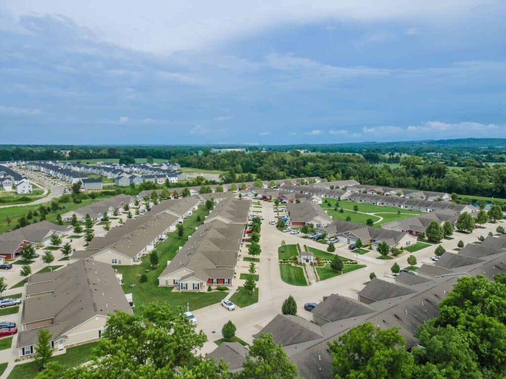 Aerial view of a suburban residential neighborhood with rows of houses, neatly arranged streets, green lawns, and scattered trees under a partly cloudy sky.