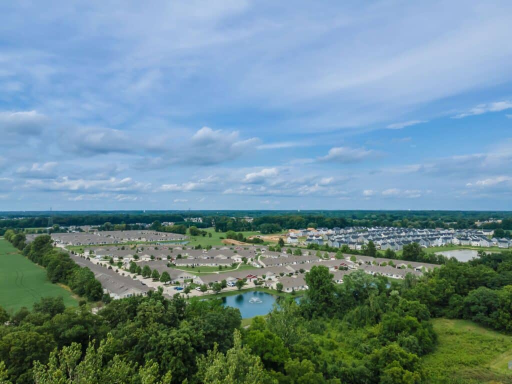 Aerial view of a suburban neighborhood with houses, small lakes, and surrounding green trees under a partly cloudy sky.