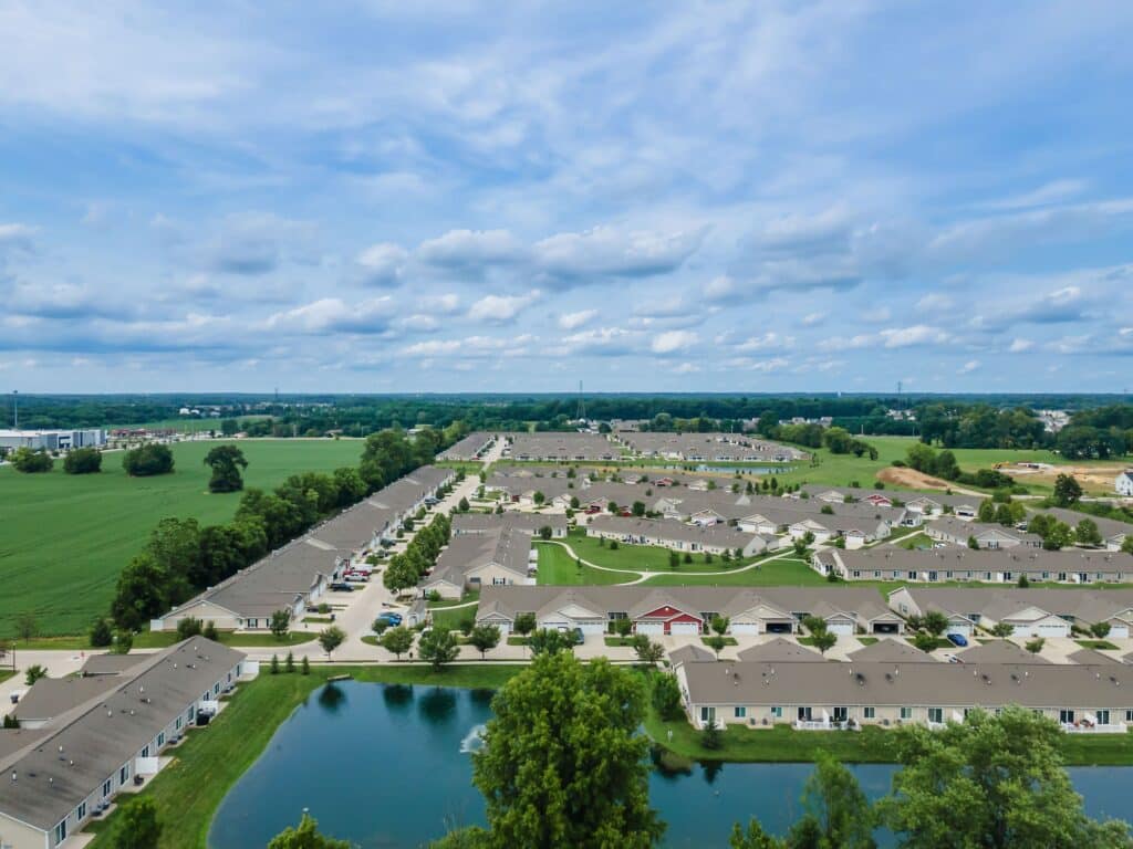 Aerial view of a suburban residential neighborhood with rows of single-story houses, green lawns, a pond, and open fields under a partly cloudy sky.