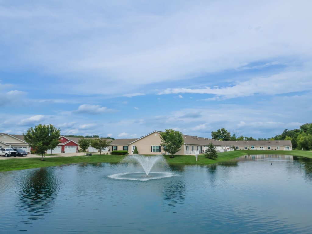 Single-story houses and trees line the far side of a pond with a central fountain, under a partly cloudy blue sky.