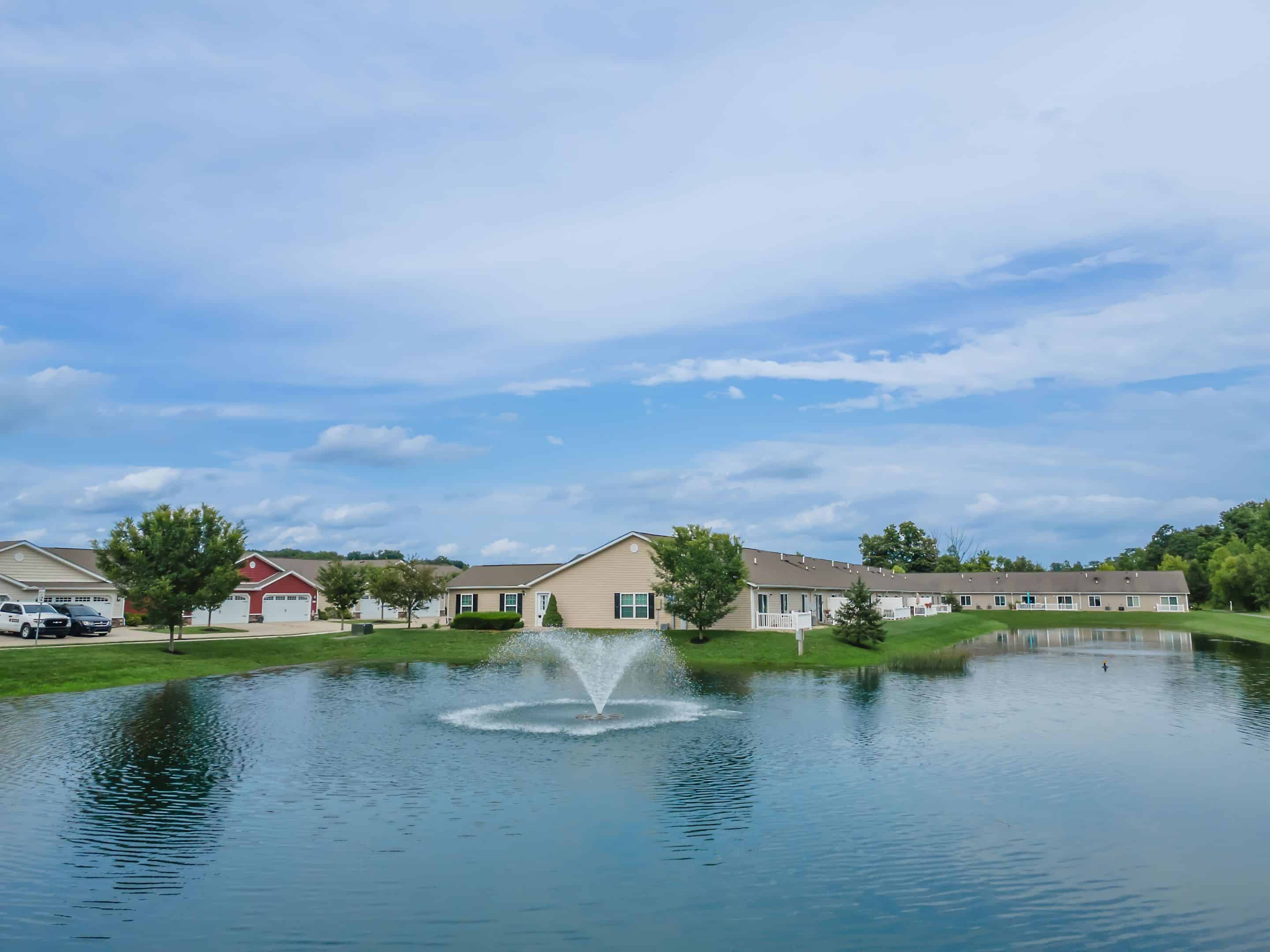 Single-story houses and trees line the far side of a pond with a central fountain, under a partly cloudy blue sky.