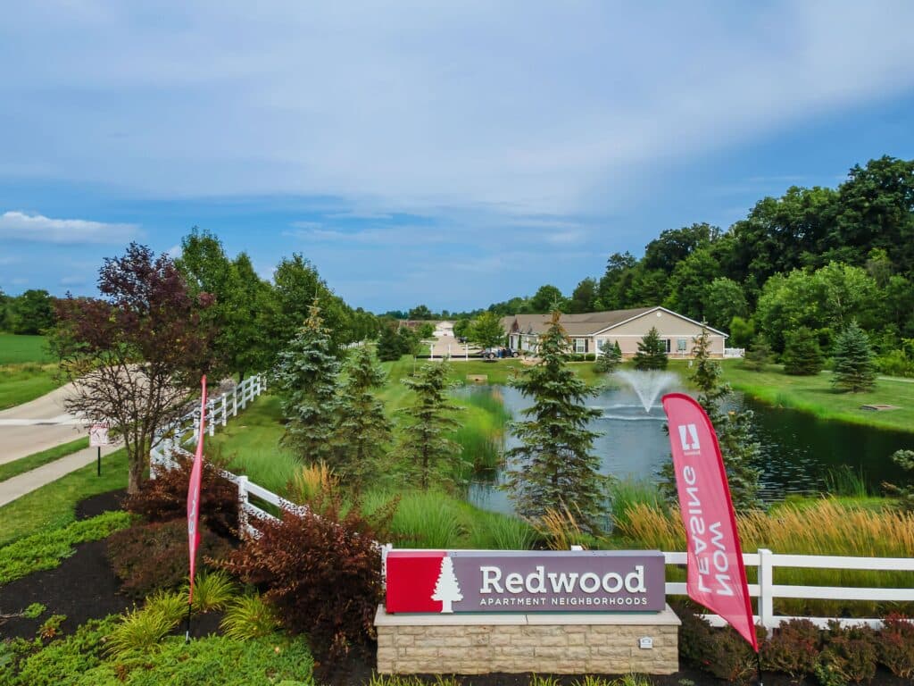 Entrance to Redwood Apartment Neighborhoods, featuring a pond, fountain, landscaping, and signage with trees and a residential building in the background.