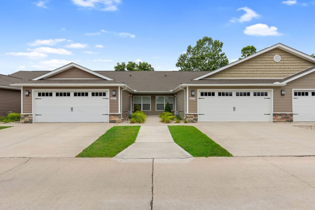 A duplex with two separate garages, beige siding, and a shared walkway, surrounded by small lawns and a concrete driveway under a blue sky.