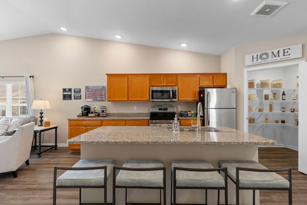 Modern kitchen with granite island, stainless steel appliances, wooden cabinets, and a walk-in pantry. Four cushioned stools are lined up at the island.