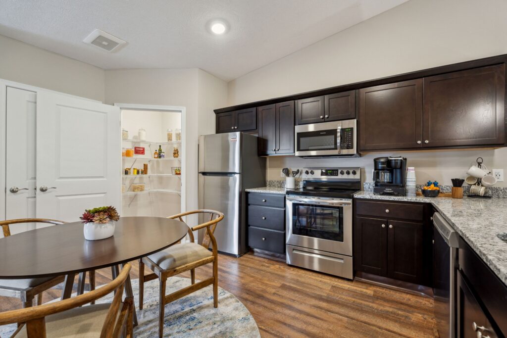 Modern kitchen with dark wood cabinets, stainless steel appliances, granite countertops, and a round dining table with four chairs; open pantry visible in the background.