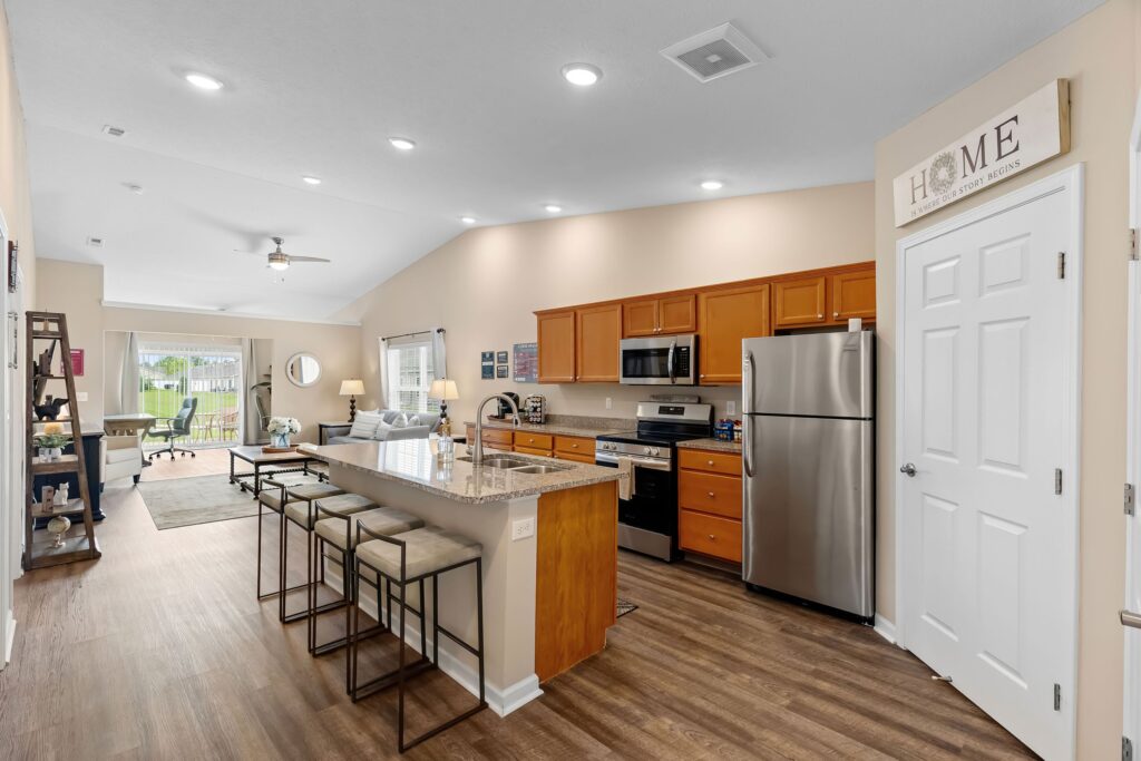 Modern kitchen with stainless steel appliances, wooden cabinets, a granite island with four barstools, and an open view to a living area with large windows.