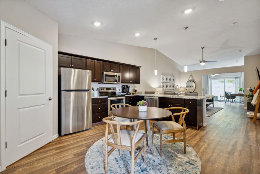 Modern kitchen and dining area with dark wood cabinets, stainless steel appliances, a round table with four chairs, and an open layout leading to a living space with natural light.