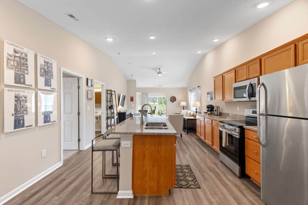 Modern kitchen and living area with wood cabinets, stainless steel appliances, island with seating, and open layout leading to a bright living room with a window.