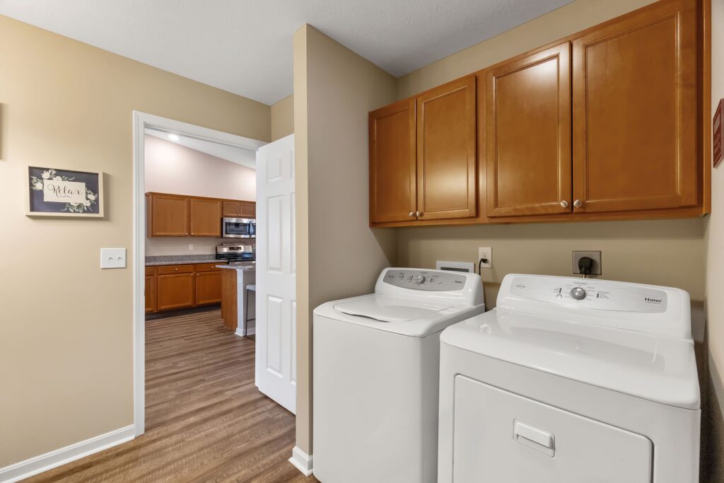 Laundry room with a washing machine and dryer under wooden cabinets, adjacent to a kitchen area with similar cabinetry and wood flooring.
